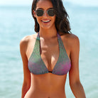 This is a tight-cropped photo of a model showing only the front of her swimsuit top. The model has mid-length black hair and black gold-rimmed sunglasses, and the ocean is out of focus in the background. The model's arms are by her side, and she is smiling into the camera. The model is wearing the Salt Skins Separates Bikini Top and Swimsuit, which has gradients of green and purple with a subtle texture pattern on the bikini top.