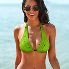 This is a tight-cropped photo of a model showing only the front of her swimsuit top. The model has mid-length black hair and black gold-rimmed sunglasses, and the ocean is out of focus in the background. The model's arms are by her side, and she is smiling into the camera. The model shows the Salt Skins Separates Collection Bikini Top and Swimsuit, which has a speckled bright green pattern and very few black dots on the bikini top.