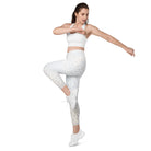 A white-skinned girl wearing a cropped workout bra with white light-patterned yoga pants with a crossover flat front waist is standing on one leg with her other leg in a yoga pose against a white wall.  She is also wearing white tennis shoes to match her athletic pants.