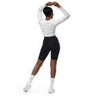 A dark-skinned girl wearing a cropped, long-sleeved workout top with a white light pattern with black workout shorts and white shoes with white socks against a white wall showing the back of her yoga top.