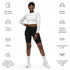 A dark-skinned girl wearing a cropped, long-sleeved workout top with a white light pattern with jeans and white shoes against a white wall showing the front of her yoga top. The image's icons display a cover-stitched collar, raglan sleeves, a tear-away label, and a fitted, double-layered waistband.