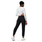 A dark-skinned girl wearing a cropped, long-sleeved workout top with a white light pattern with jeans and white shoes against a white wall showing the back of her yoga top.