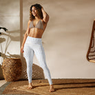 This full-length front view of a girl wearing tight white yoga leggings with beautiful subtle check patterns in a performance fabric with a tan workout bra. The leggings have a double-layered flat front waist. The brown-haired woman is standing on a wicker yoga mat with one hand by her side and the other hand combing through her hair.