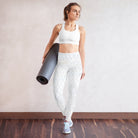 A picture of a girl standing against a white or tan wall with a yoga mat in her hands. She is wearing lovely white yoga leggings with a double-layered waist and a flat front with subtle patterns. She is also wearing light blue tennis shoes standing on a wooden floor.