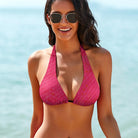 This is a tight-cropped photo of a model showing only the front of her swimsuit top. The model has mid-length black hair and black gold-rimmed sunglasses, and the ocean is out of focus in the background. The model's arms are by her side, and she is smiling into the camera. The model is wearing the Salt Skins Separates Bikini Top only. The bikini top is a hot pink color with a subtle texture pattern.